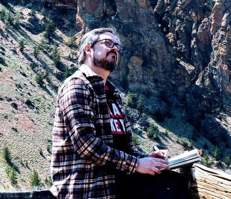 Man with glasses and a beard, wearing a plaid shirt over a graphic t-shirt, sitting outdoors in front of a rocky mountain landscape with sparse trees, holding a notebook and pen.